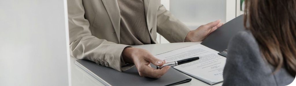 Zwei Personen sitzen an einem Tisch, zwischen ihnen liegt eine Mappe und es wird mit dem Stift auf Text in der Mappe gedeutet. Man sieht keine Gesichter, nur den Oberkörper und die Hände.